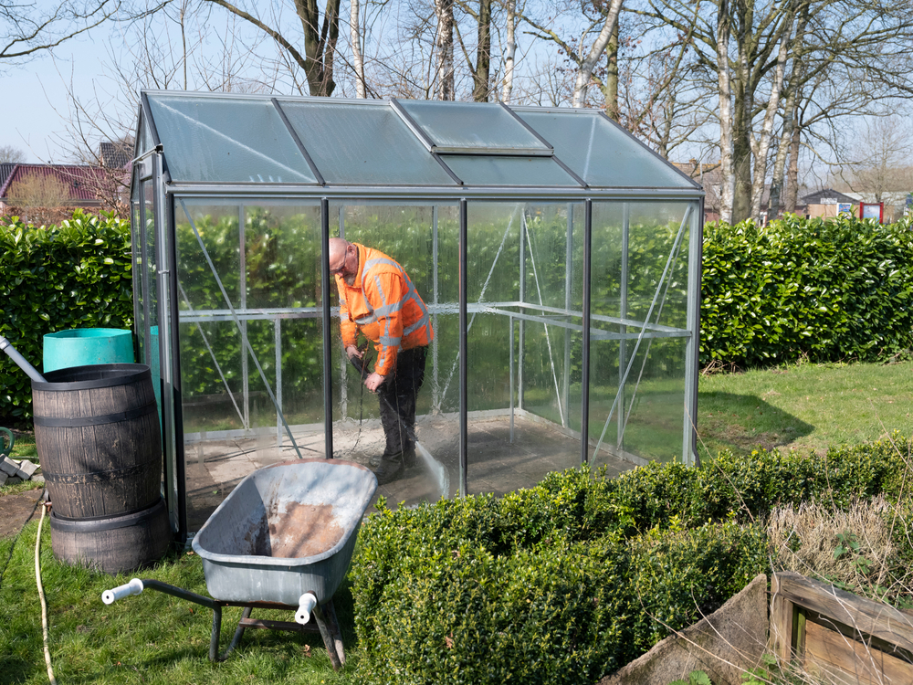Construction worker cleaning filth with high pressure cleaner from a glass greenhouse.